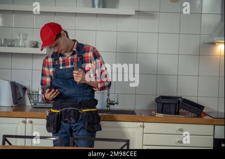 Un tecnico di riparazione serio e attento si trova al bancone della cucina e guarda lo schermo dello smartphone Foto Stock