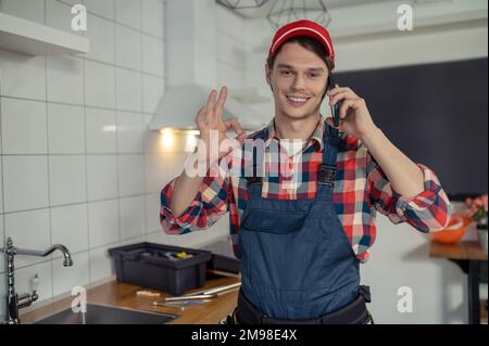 Ritratto in vita di un soddisfatto tecnico idraulico che fa il segno OK durante la telefonata in una cucina cliente Foto Stock