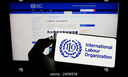 Persona che tiene il cellulare con il logo dell'agenzia ONU International Labour Organization (ILO) sullo schermo di fronte alla pagina web. Messa a fuoco sul display del telefono. Foto Stock