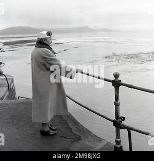 1950s, storica, anziana donna che indossa un lungo cappotto, con colletto in pelliccia, guanti e cappello decorati con piume, in piedi su una ringhiera di un porto, che si affaccia su una baia..... quali ricordi di vita? Inghilterra, Regno Unito. Foto Stock