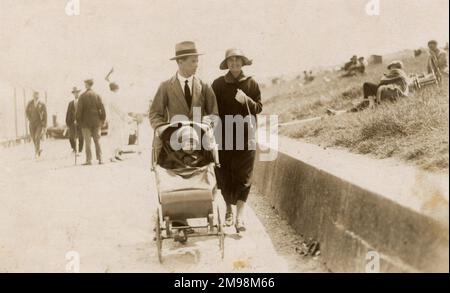 Una coppia intelligente passeggiando lungo il lungomare con il bambino in buggy, avvolto contro la (probabile) brezza marina! Foto Stock