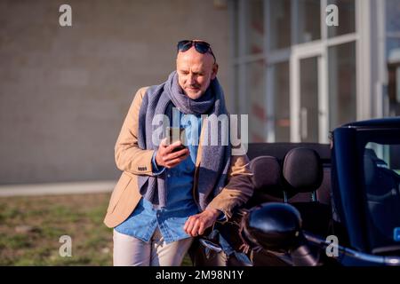 Immagine di un uomo di mezza età che indossa abiti casual e utilizza il telefono cellulare mentre si è in piedi fuori accanto alla vettura hia. Foto Stock