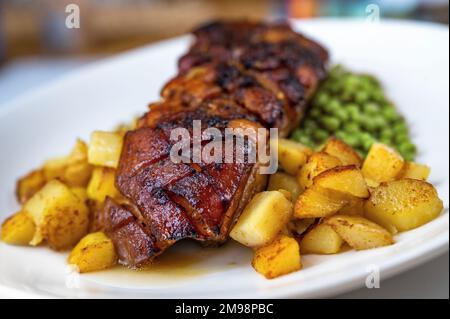 Pancia di maiale fritta al forno con pelle croccante e patate e piselli su piatto bianco sul tavolo, primo piano. Foto Stock