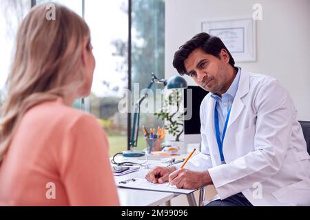 Medico di sesso maschile o GP Meeting paziente di sesso femminile maturo per appuntamento in ufficio facendo note sulla clipboard Foto Stock