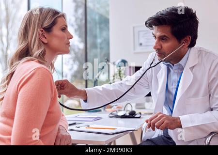 Medico o GP di sesso maschile con mantello bianco che esamina la donna matura che ascolta il torace con stetoscopio Foto Stock