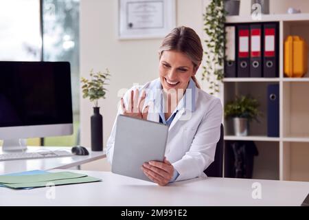 Dottore o GP che indossa un cappotto bianco seduto alla scrivania in ufficio con videochiamata sul tablet digitale Foto Stock