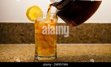 Versare il tè freddo appena preparato in un bicchiere trasparente. Caraffa di tè versando in un bicchiere di ghiaccio con una fetta di limone. Foto Stock