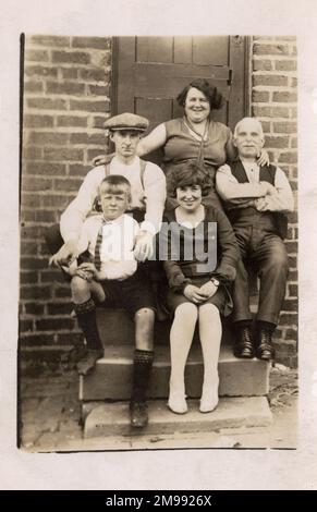 La famiglia d'inverno in un giorno d'estate! In posa per un ritratto di gruppo di famiglia sul gradino posteriore della loro casa. Tre generazioni nella foto. Foto Stock