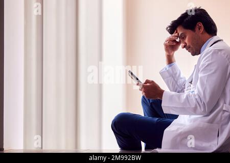 Stressato medico di sesso maschile indossando camice bianco seduto sul pavimento in corridoio di ospedale con telefono cellulare Foto Stock
