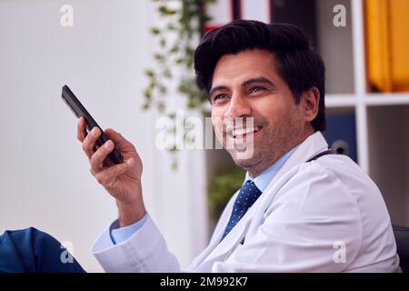 Sorridente medico maschile o GP indossare camice bianco seduto alla scrivania in ufficio con telefono cellulare Foto Stock