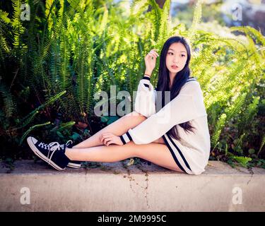 Bella giovane donna in abito maglione corto seduto sul muro con Ferns dietro di lei Foto Stock
