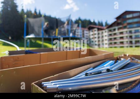 Grandi scatole di cartone con travi in ferro e altro kit per assemblare un trampolino a rete come sullo sfondo accanto a persone che assemblano un trampolino su un b Foto Stock