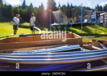 Grandi scatole di cartone con travi in ferro e altro kit per assemblare un trampolino a rete come sullo sfondo accanto a persone che assemblano un trampolino su un b Foto Stock