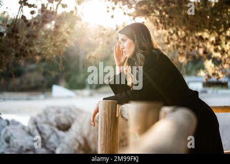 Donna triste vestita di nero guardando il mare con il tramonto sullo sfondo Foto Stock