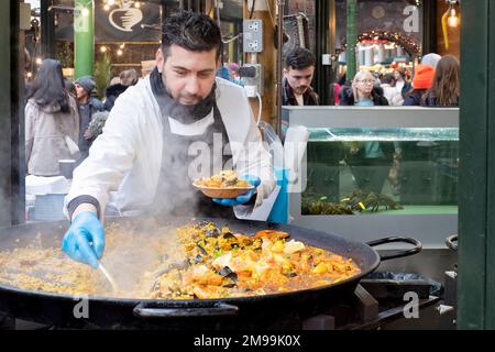 Uno chef che sceglie un piatto da asporto che serve da una grande paella di pesce dalla bancarella del mercato alimentare Furness Fish Grill a Borough Market, Londra Foto Stock