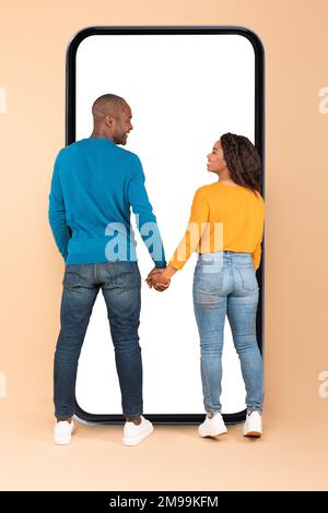 Felice coppia nera in piedi vicino al telefono enorme con schermo vuoto, in piedi di nuovo alla macchina fotografica, tenendo le mani e sorridendo Foto Stock