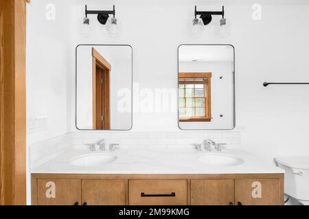 Un bel bagno con un armadietto di legno, banco di marmo, backsplash della metropolitana e luci nere sopra gli specchi. Foto Stock