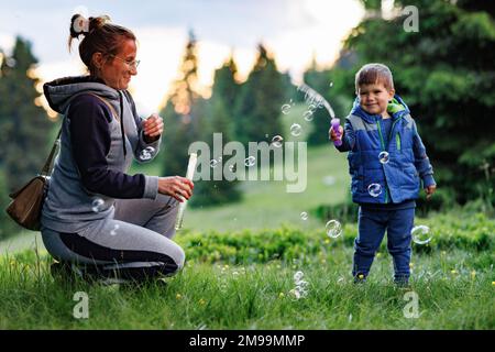 Una felice madre sorridente premurosa in un vestito sportivo grigio caldo gioca con il suo piccolo figlio allegro in un vestito blu carino su un prato verde e gli insegna a blo Foto Stock