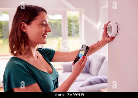 Donna che utilizza l'app sul telefono cellulare per controllare il termostato di riscaldamento centrale a casa Foto Stock