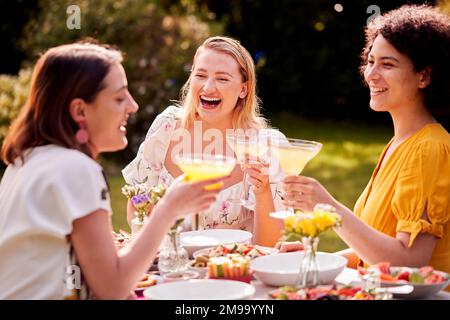 Tre amici femminili seduti all'aperto nel Giardino d'Estate a casa bere cocktail e mangiare pasto Foto Stock