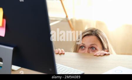 Donna si nasconde dietro la scrivania dell'ufficio e guarda il monitor del computer. Foto Stock