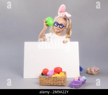 Immagine del concetto di Pasqua. Bambina con occhiali, con orecchie conigliate, curiosamente guardando l'uovo. Capretto nascosto dietro lavagna bianca su sfondo grigio ne Foto Stock