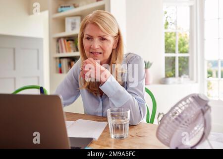 Donna mature menopausale che ha il flusso caldo a casa di raffreddamento se stessa con ventilatore collegato al laptop Foto Stock