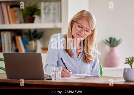 Donna matura che lavora sul laptop seduto al tavolo a casa Foto Stock