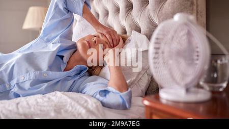 Donna matura in menopausa che soffre di insonnia a letto a casa utilizzando ventilatore elettrico Foto Stock