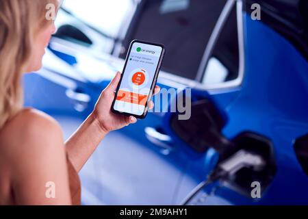 Donna che carica l'auto elettrica con il cavo utilizzando l'app sul telefono per monitorare il livello della batteria Foto Stock