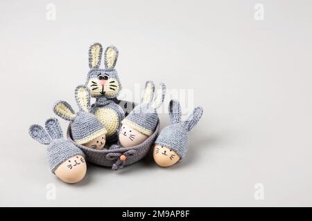 Cinque uova di Pasqua con volti divertenti e cappelli grigi uncinati con orecchie conigliate in un cestino decorativo e un coniglietto grigio uncinato su uno sfondo grigio. Happ Foto Stock