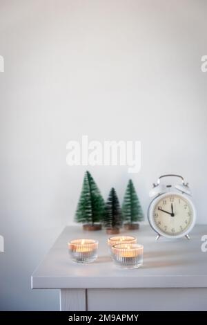 Una sveglia bianca, un puntatore a dodici, un piccolo albero di Natale artificiale verde e candele accese in candelabri su una cassettiera bianca. Copia Foto Stock