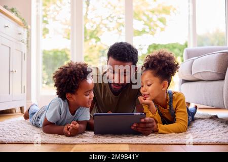 Padre a casa sdraiato su Rug in Lounge con i bambini che utilizzano il tablet digitale Foto Stock