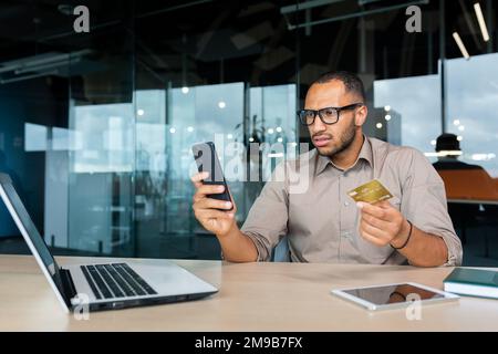 Insoddisfatto e arrabbiato uomo d'affari in ufficio cercando di fare l'acquisto in negozio online, uomo ispanico tenendo la carta di credito e smartphone in mano, l'uomo truffato ricevuto errore frode. Foto Stock