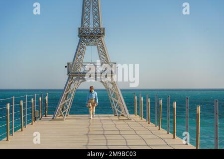 Grande modello della Torre Eiffel sulla spiaggia. Una donna cammina lungo il molo verso la torre, indossando una giacca blu e jeans bianchi. Foto Stock