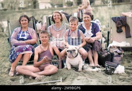 Sulla spiaggia di Margate: Tre donne si siedono in sedie a sdraio, mentre due ragazzi in costume da bagno siedono, uno sulla sabbia, uno a cavallo di un bulldog giocattolo Foto Stock