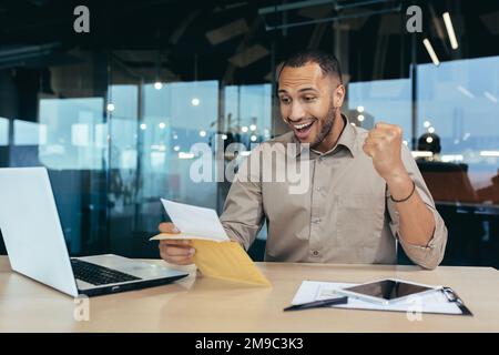 Un uomo d'affari di successo ha ricevuto una lettera di notifica con buone notizie, l'uomo all'interno dell'ufficio apre e legge la busta della posta con il messaggio celebra il successo della vittoria, l'uomo ispanico tiene la mano in su il gesto trionfale. Foto Stock