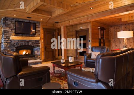 Divani e poltrona in pelle marrone di fronte al caminetto illuminato in pietra naturale nella camera per famiglie al piano terra, all'interno di un profilo piatto in legno e di una casa in legno. Foto Stock