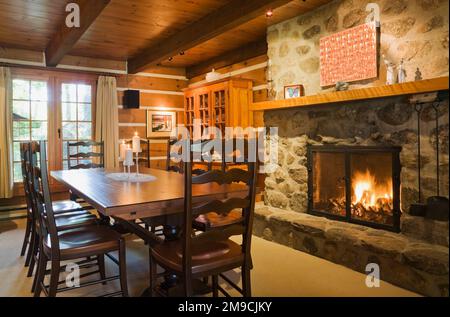 Lungo tavolo da pranzo in legno con sedie a schienale alto accanto al caminetto a legna illuminato in pietra di campo nella sala da pranzo all'interno di una casa in legno in stile canadese. Foto Stock