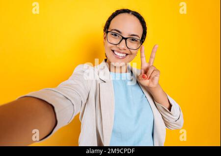 Bella, carismatica positiva ispanica o brasiliana con gli occhiali, scatta una foto selfie sul suo smartphone, guarda la fotocamera, sorride, mostra segno di pace con le dita, sfondo giallo isolato Foto Stock