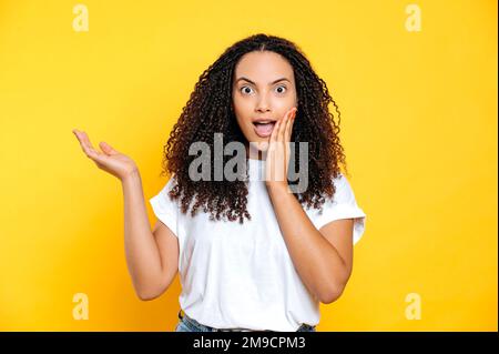 Sorpresa scioccata ispanica o brasiliana donna, guarda la fotocamera in stupore con la bocca leggermente aperta, con la mano punta a lato in uno spazio vuoto, si trova su sfondo giallo isolato Foto Stock