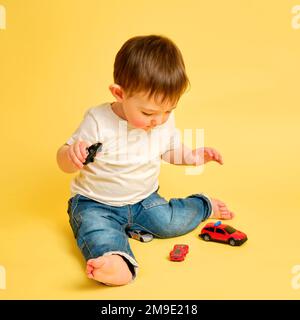 Il bambino del bambino gioca con le automobili del giocattolo su uno sfondo giallo dello studio. Felice bambino in t-shirt bianca e jeans blu gioca con le auto giocattolo. Bambino di un anno e oltre Foto Stock