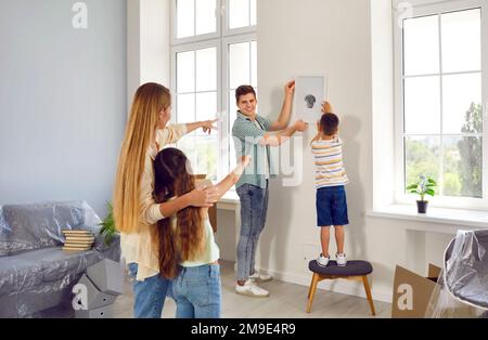 L'uomo gioioso e il suo figliolo appendono insieme un quadro sul muro sotto la guida di una donna e di una figlia. Foto Stock