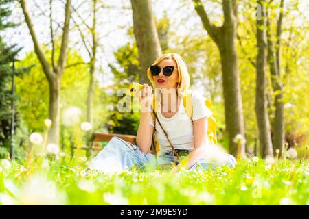 Una donna bionda turistica che invia una nota vocale con il telefono, indossando un cappello e occhiali da sole seduti sull'erba in primavera accanto alle margherite in un parco in Foto Stock