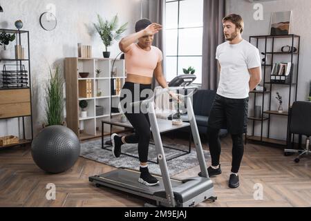 Home fitness allenamento sportivo donna e uomo allenamento sulla pista da corsa. Ritratto di una giovane donna africana focalizzata che indossa abbigliamento sportivo e tapis roulant e il suo allenatore uomo caucasico. Foto Stock