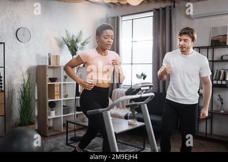 Home fitness allenamento sportivo donna e uomo allenamento sulla pista da corsa. Ritratto di una giovane donna africana focalizzata che indossa abbigliamento sportivo e tapis roulant e il suo allenatore uomo caucasico. Foto Stock