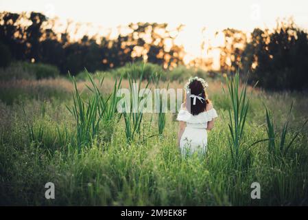 ragazza in abito bianco con una corona floreale risalta sul campo in estate al tramonto Foto Stock