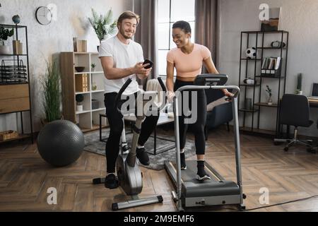 Felice coppia sorridente che fa cardio sulla bici stazionaria e sul tapis roulant usando il telefono astuto per guardare le video lezioni in linea come il peso unico. Attraente femmina e maschio allenarsi sul fitness a casa. Foto Stock