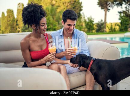 Una giovane coppia interracial si siede su un divano da giardino in una lussuosa villa o resort. Tenere bicchieri di succo d'arancia e stanno sorridendo mentre coccolare un cane - D Foto Stock