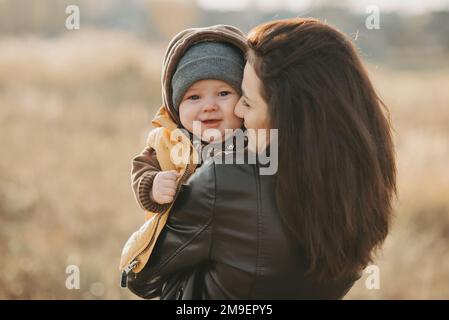 Felice madre e figlio. La giovane ragazza che ride abbraccia il suo bambino tra le braccia in autunno all'aperto Foto Stock